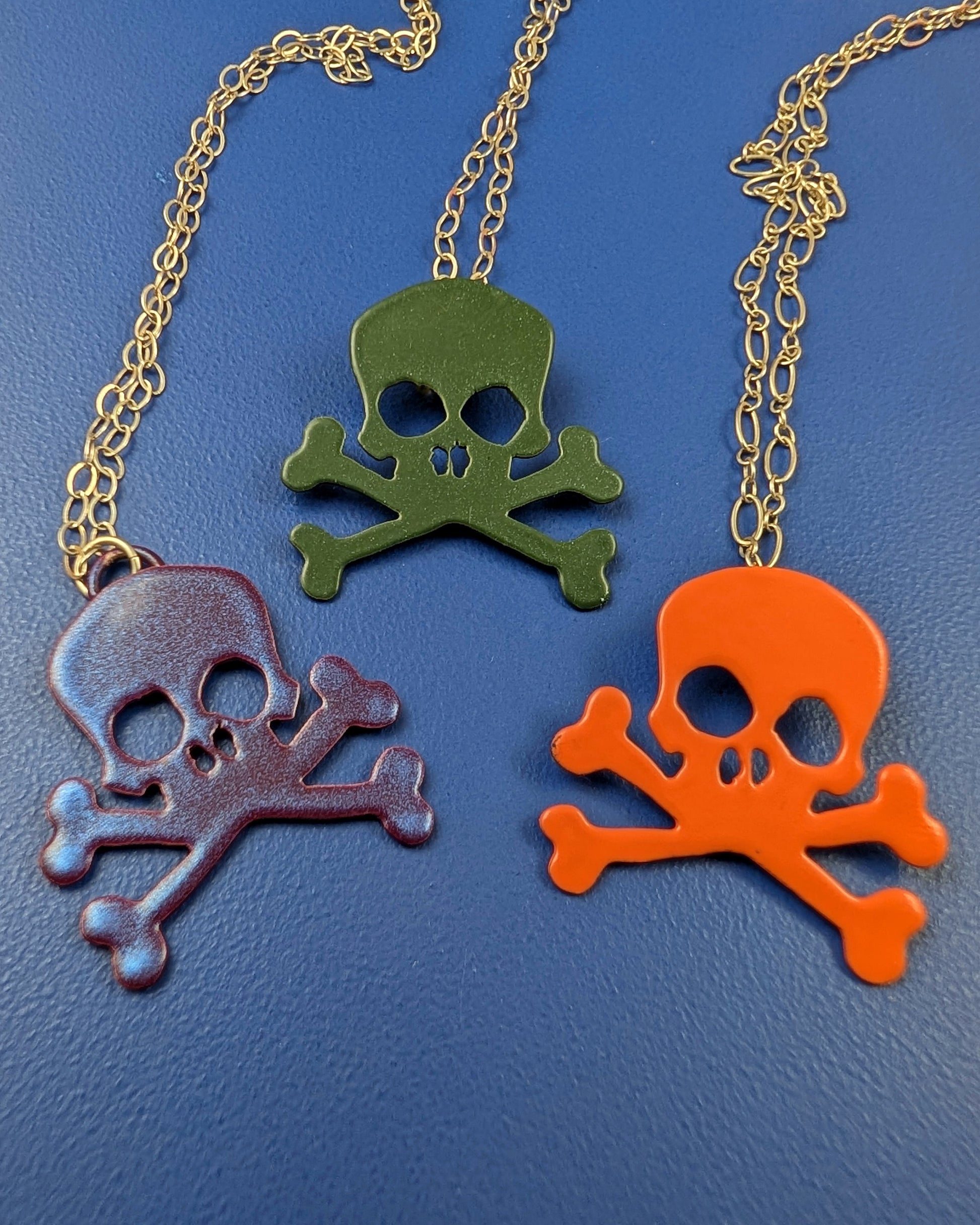 Three skull and crossbones pendants on silver chains against a blue background.