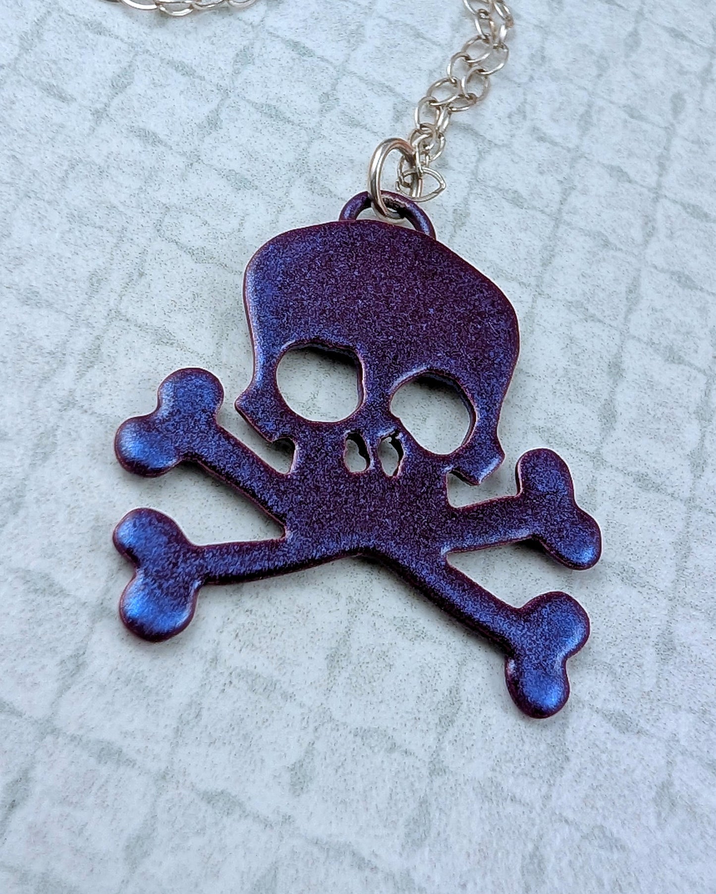 Purple skull and crossbones pendant on a chain against a light gray textured background