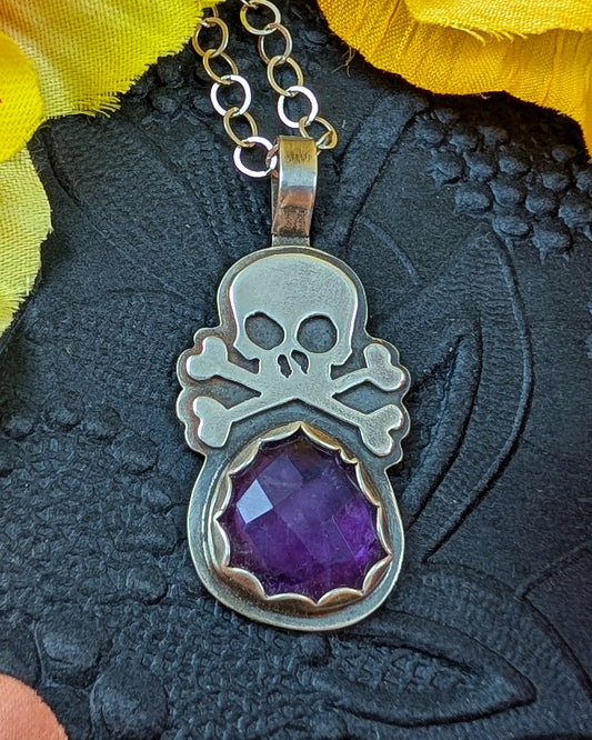 Silver skull and crossbones pendant with a purple amethyst gemstone on a textured black surface.