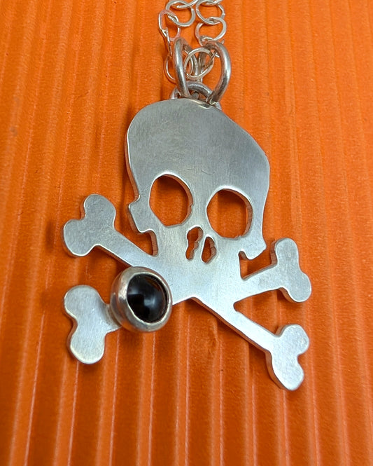 Silver skull and crossbones pendant with a black gemstone on an orange background