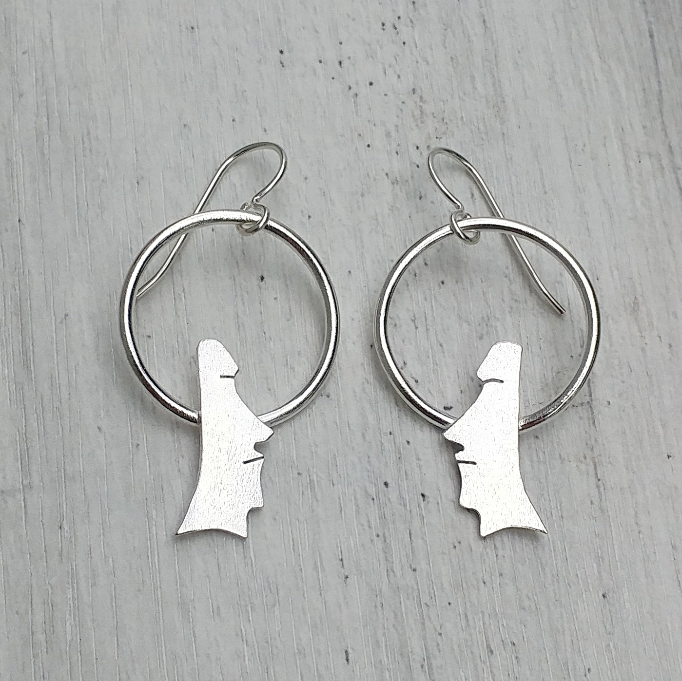 Another view of Handmade sterling silver moai head earrings on circles on a grey background.