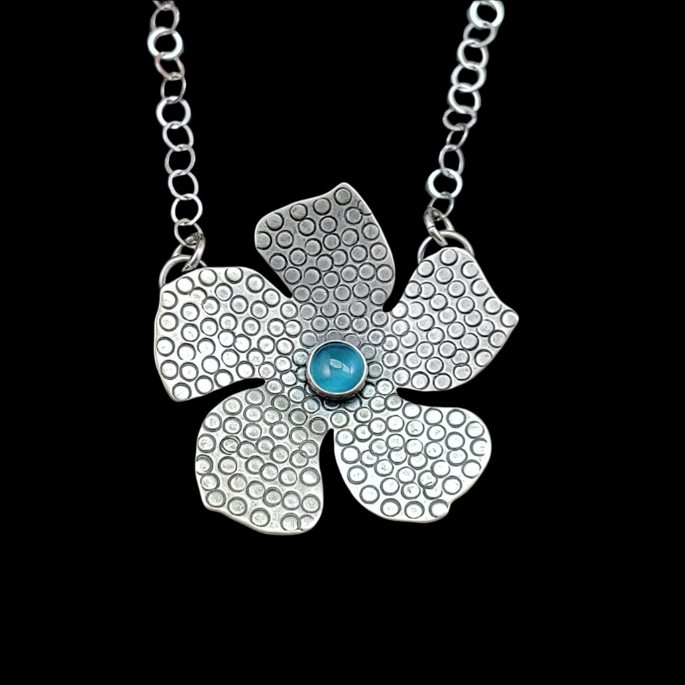 Handmade tiki flower in sterling silver with round stamped details and blue stone on black background