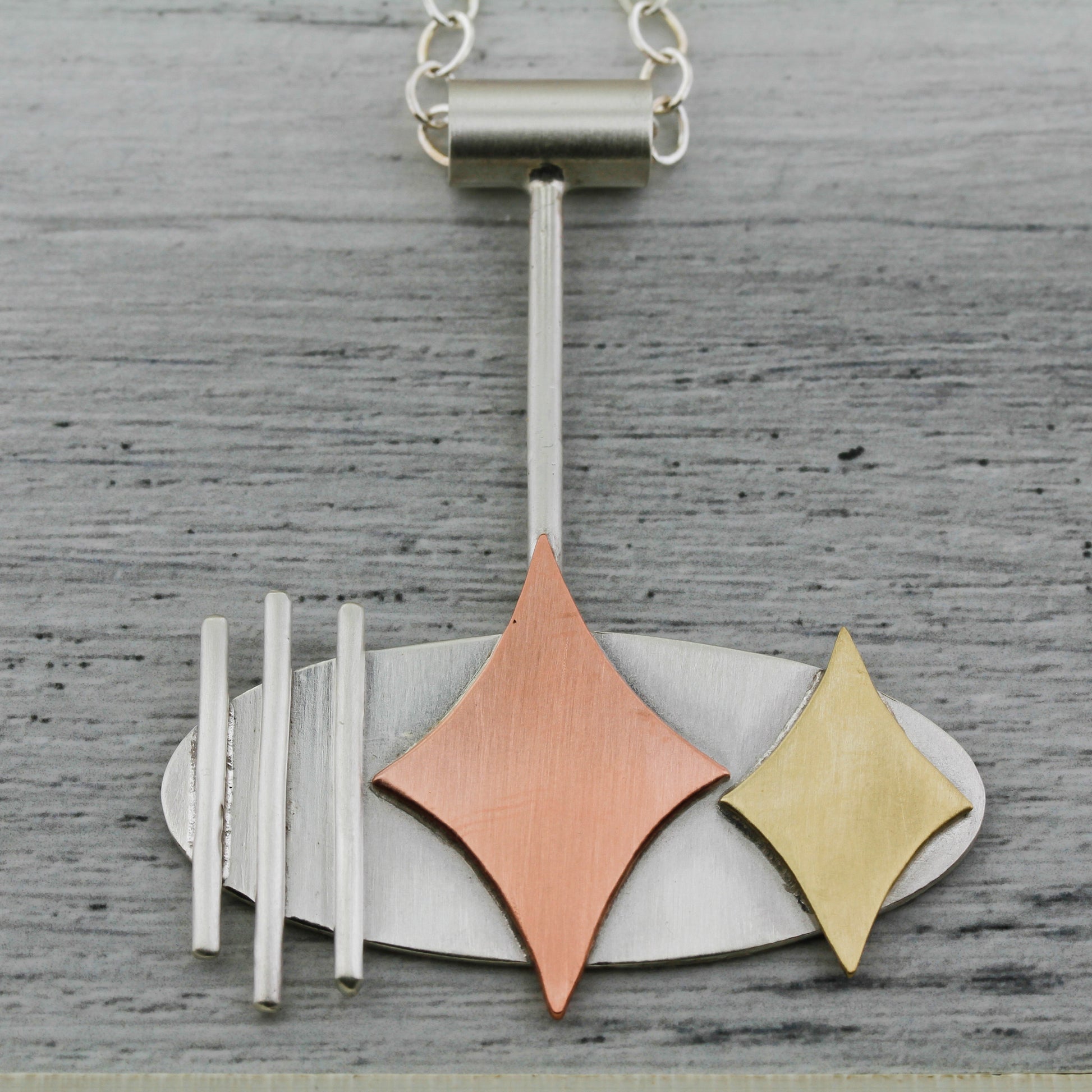 Mid century modern necklace handmade in sterling silver with copper and brass retro stars, and silver bars in an oval setting shown on a gray background