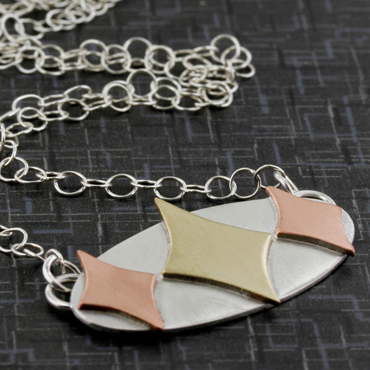 Mid century modern necklace handmade in sterling silver with copper and brass retro stars in an oval setting shown sideways on a black background