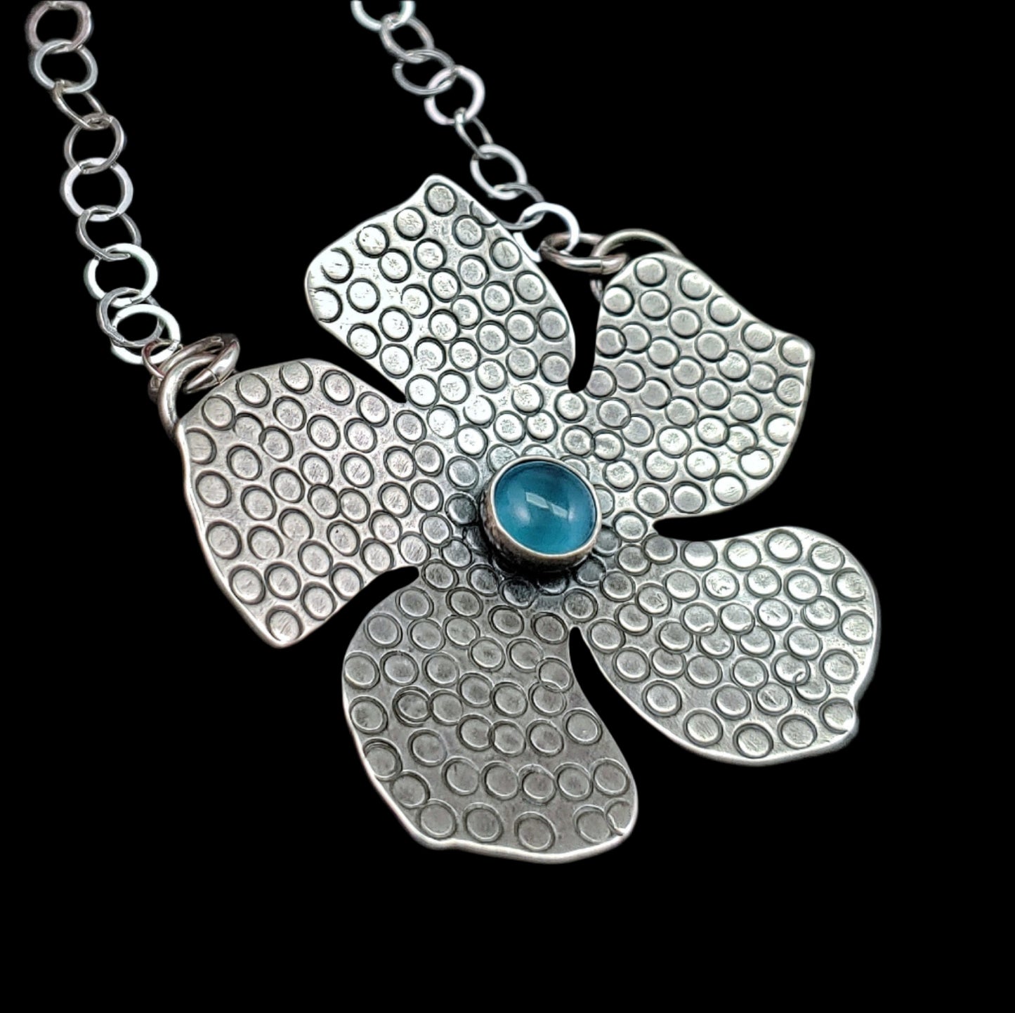 Handmade tiki flower in sterling silver with round stamped details and blue stone on a black background in a sideways position