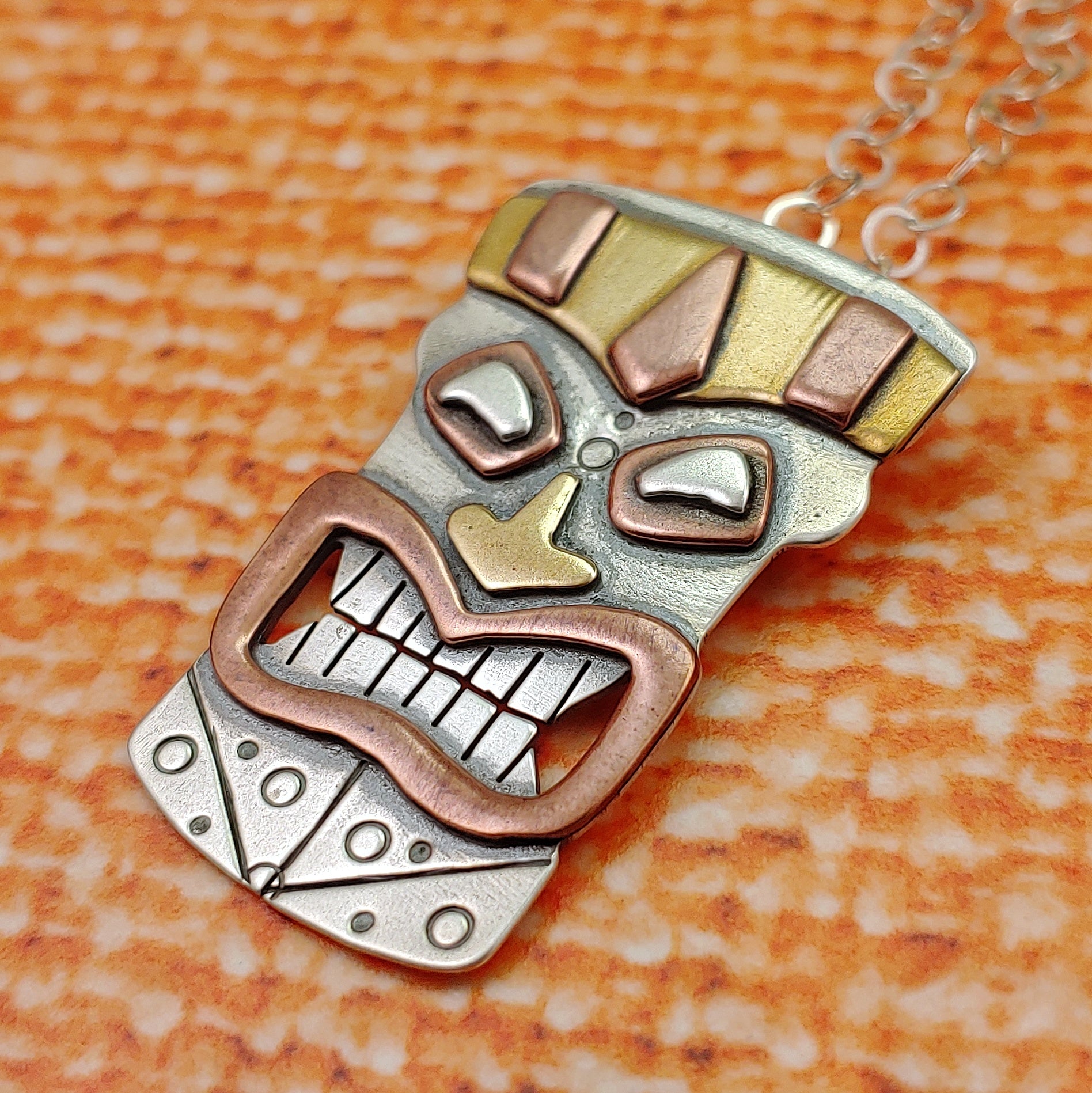 Tiki head mask necklace handmade in sterling silver, copper and brass perfect for mens tiki jewelry shown on an orange background