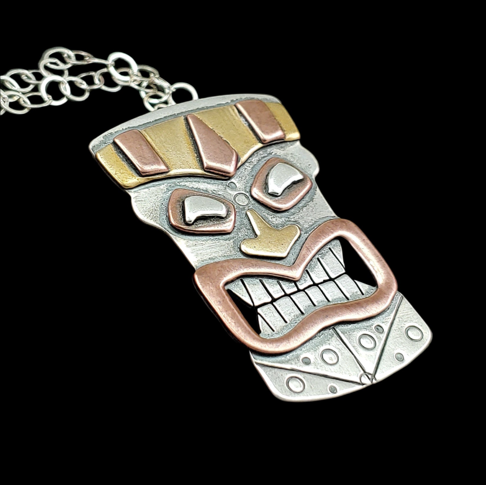 Tiki head mask necklace handmade in sterling silver, copper and brass perfect for mens tiki jewelry shown as a side angle on a black background