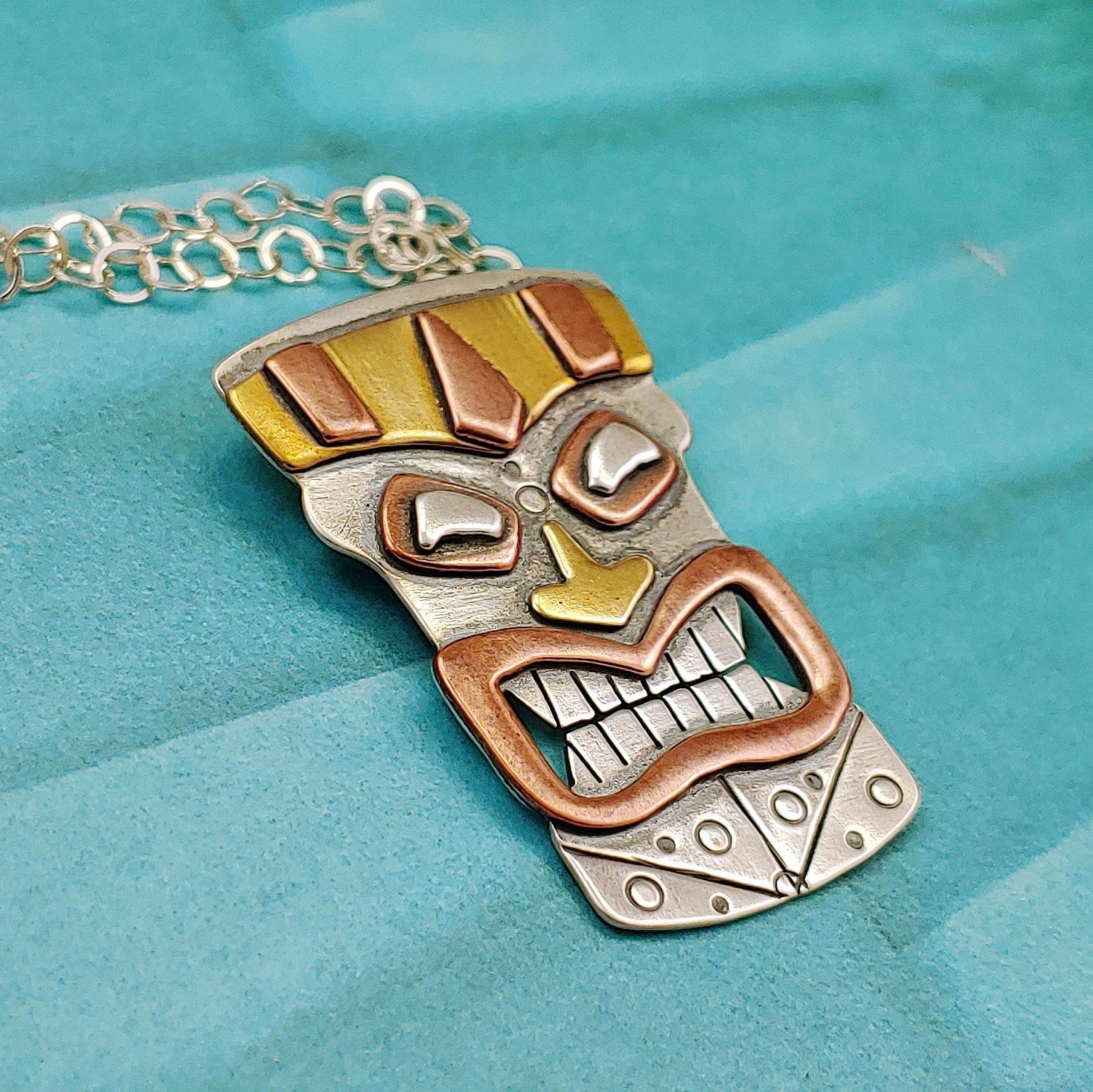 Tiki head mask necklace handmade in sterling silver, copper and brass perfect for mens tiki necklace shown on a turquoise background