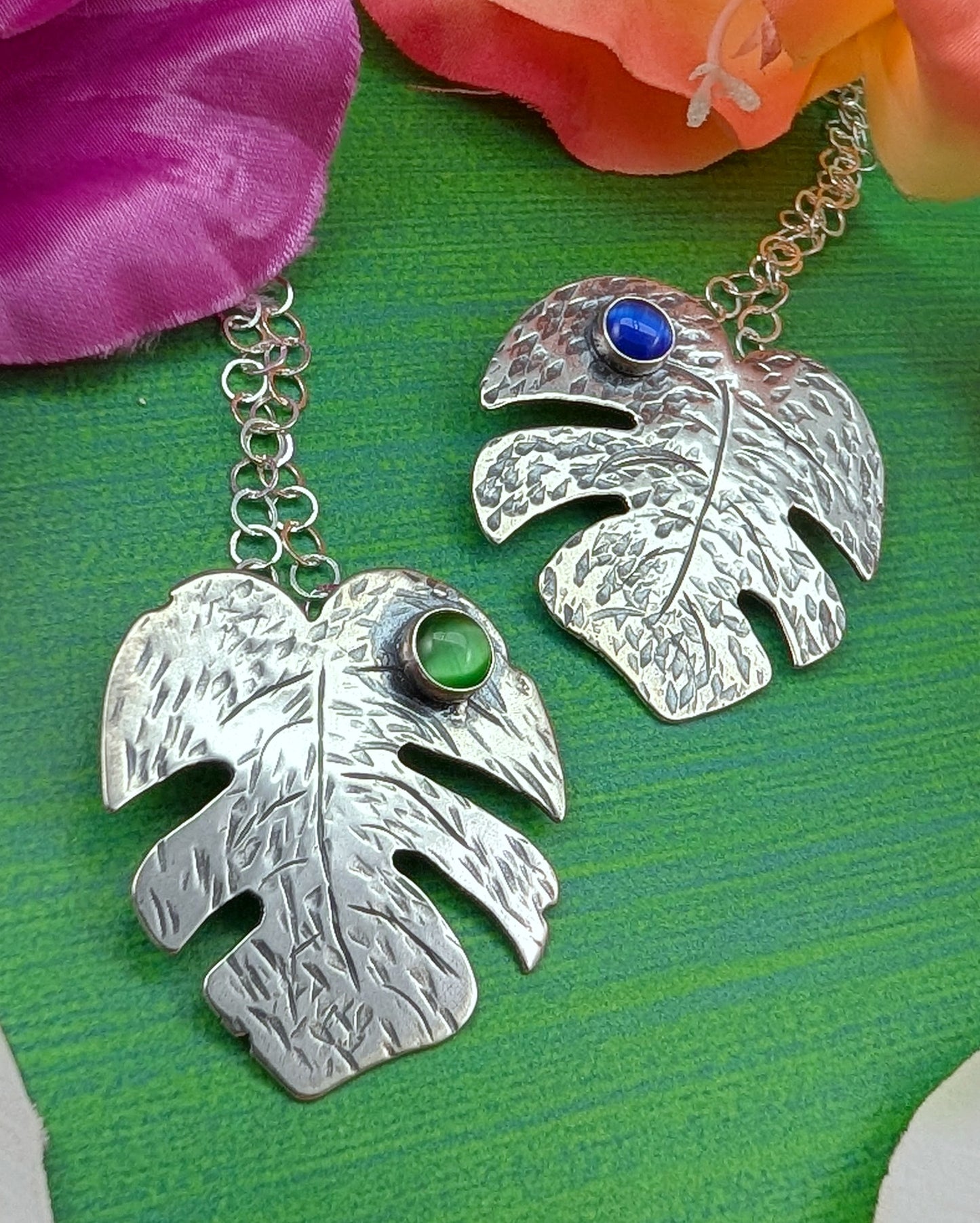Sterling silver monstera leaf necklaces with green cats eye and blue cats eye on a green background with flowers.