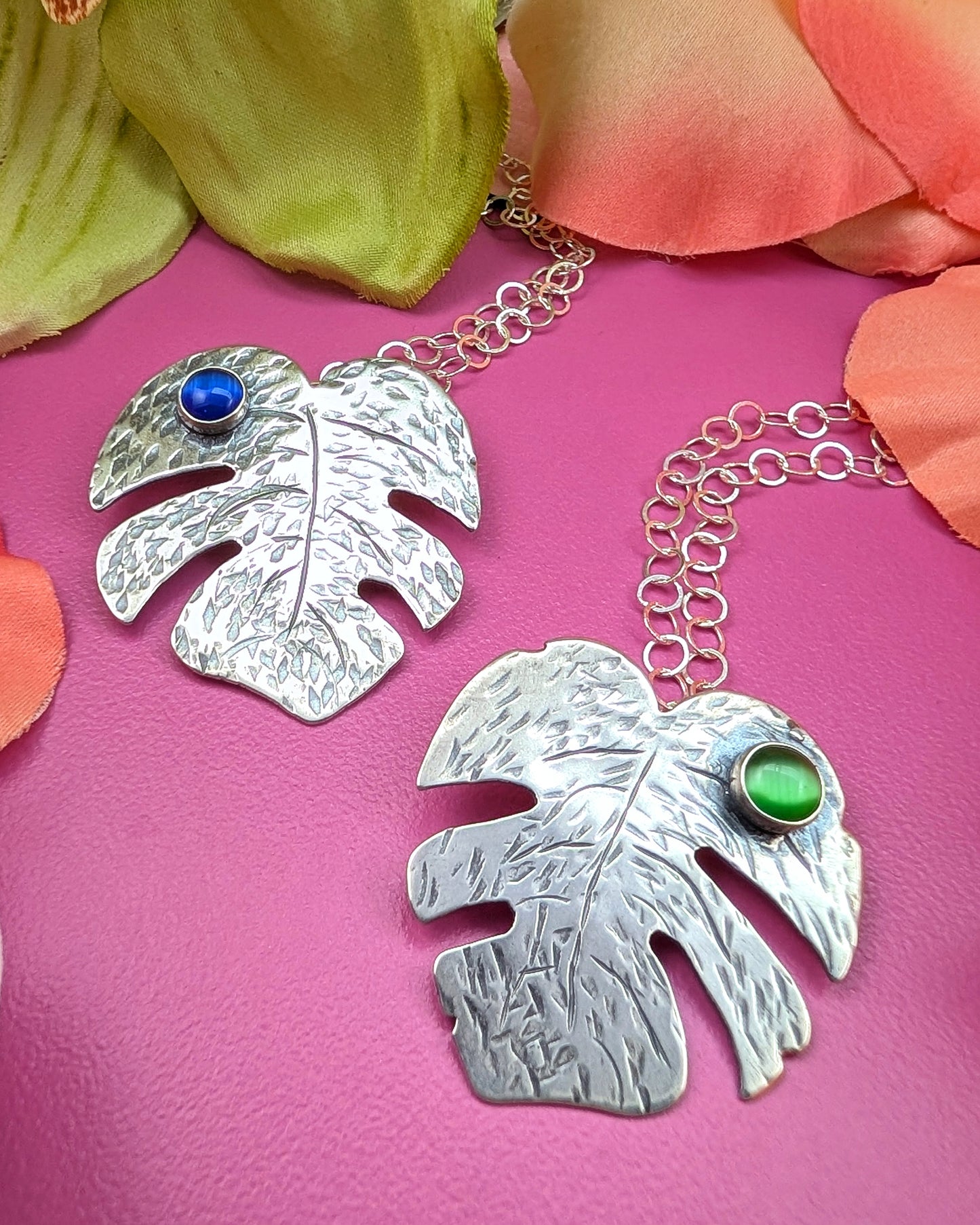 Sterling silver monstera leaf necklace swith blue cat's eye and green cat's eye on a pink background