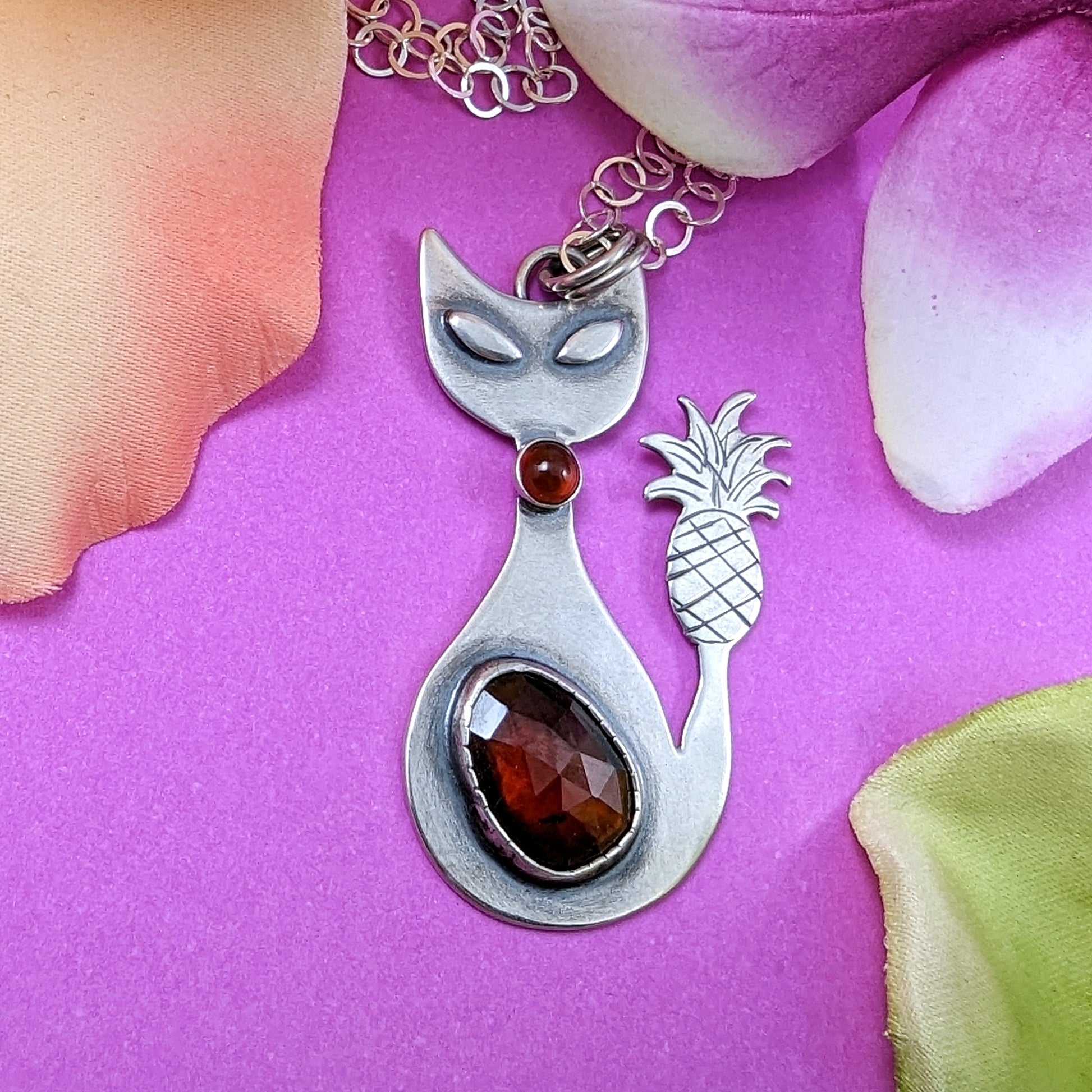 Sterling silver handmade cat with pineapple tail and garnet on pink background with flowers