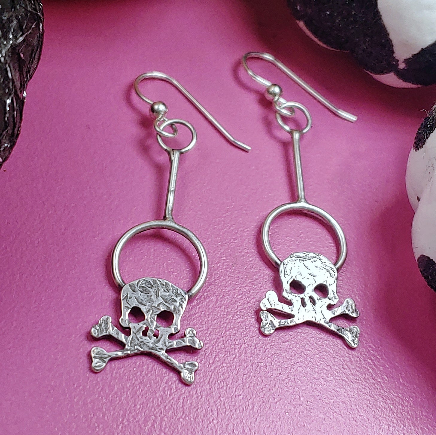 Sterling silver hammered textured skull dangle earrings on a pink background.